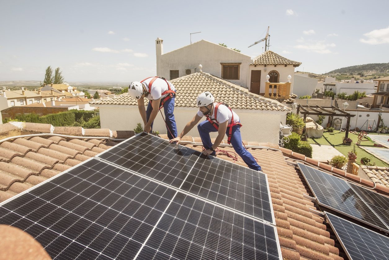Due installatori con imbracatura posano un modulo fotovoltaico su un tetto in coppi di una villa