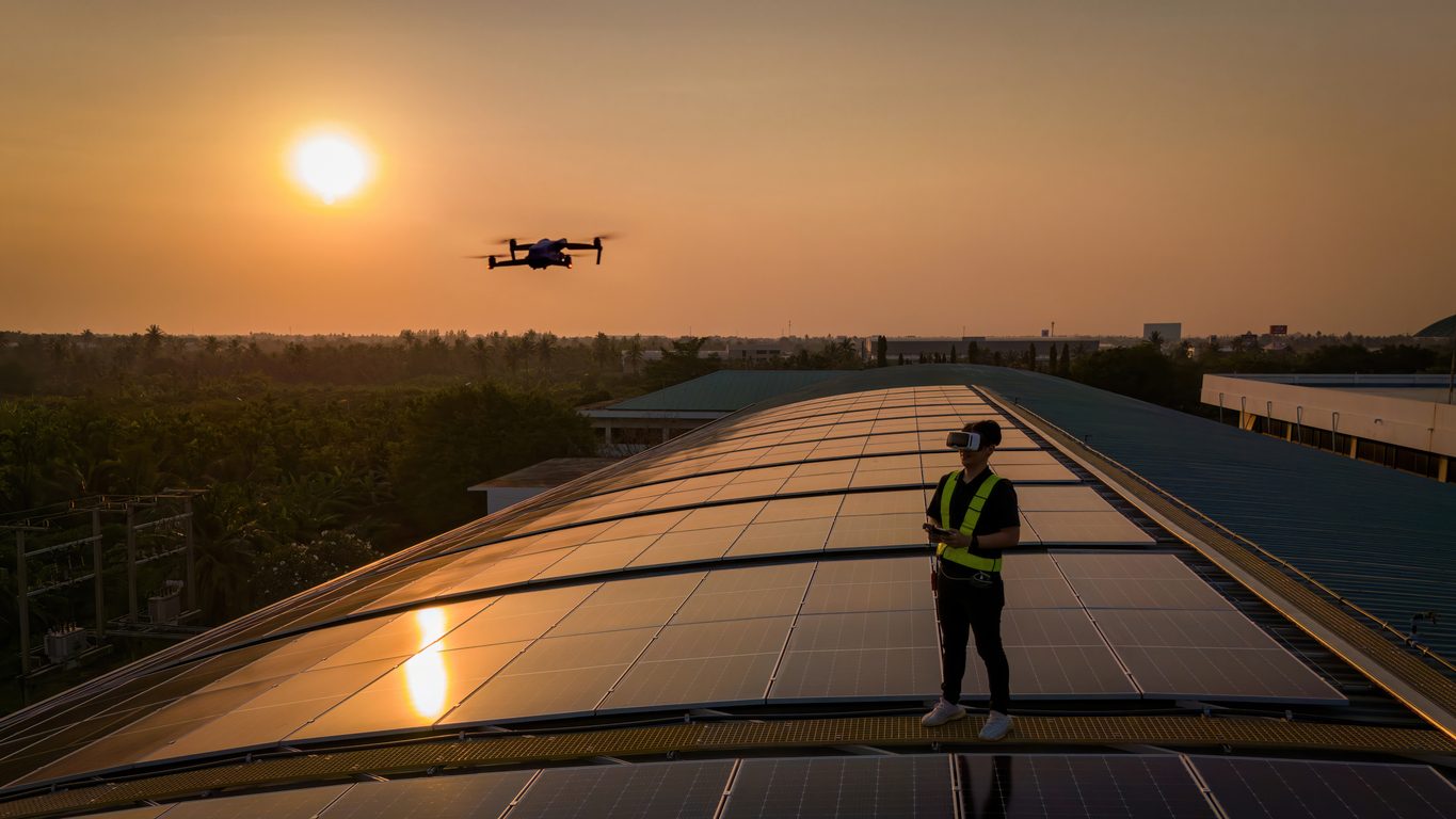 Tecnico pilota un drone per ispezionare un impianto fotovoltaico su copertura industriale al tramonto