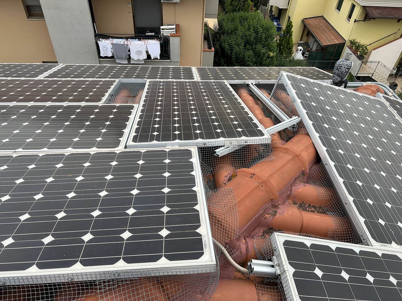 Moduli fotovoltaici montati con inclinazioni diverse a cavallo del colmo di un tetto in coppi, binari sporgenti e rete anti-piccioni aperta
