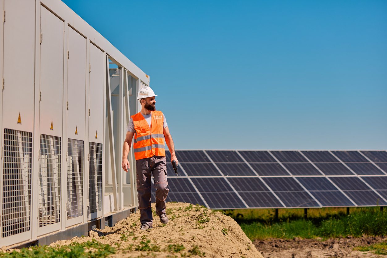 Tecnico con casco e gilet STILCA Energy cammina tra la cabina di conversione e le file di moduli di un impianto fotovoltaico a terra